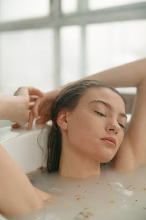 Woman relaxing in a bathtub with eyes closed, surrounded by candles and bath products, enjoying a calming self-care ritual at home